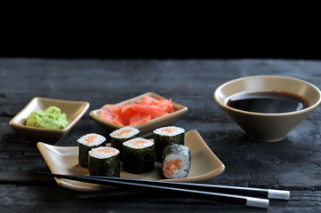 Roll with salmon on a light ceramic plate. Next to a plate of chopsticks. Marinated ginger, soy sauce and wasabi in bowls. Black wooden background. Close-up.