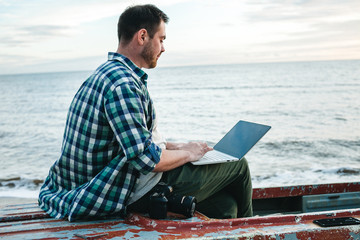 Young blogger photographer working with a laptop while sitting in an old boat on the ocean coast