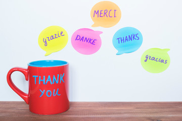 Red cup and words Thank You translated in different languages on the wooden table.