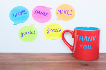 Red cup and words Thank You translated in different languages on the wooden table.