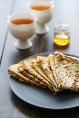 Crepes on a grey ceramic plate served with honey and 2 cups of tea. Dark wooden table, high resolution