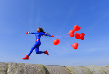 A young woman dressed in shiny blue runs and jumps  across hay bales with a bunch of red heart balloons in  her hand.