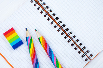 Open copybook with two rainbow-coloured pencils and a rubber on white wooden table