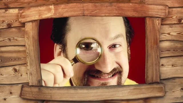 Seen from the window of a wooden cabin: a funny ugly man looking at the camera with a magnifying lens (detective, private eye), searching for a clue and finding it.