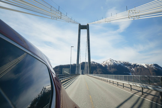 View back from side of car, driving on modern bridge at sunny day with beautiful blue sky and mountains at background. Travelling in Norway.
