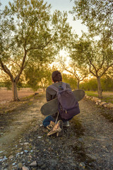 Young boy crouching and traveling for a rural path with a skateboard and a sneakers hanging of his backpack. Guy resting and looking the sunset.