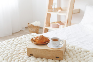 Stylish cozy scandinavian bedroom interior: bed with white linen, light beige thick yarn knitted woolen merino chunky blanket, wooden rack, tray with a croissant and a cup of tea, white wall.