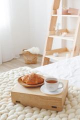 Stylish cozy scandinavian bedroom interior: bed with white linen, light beige thick yarn knitted woolen merino chunky blanket, wooden rack, tray with a croissant and a cup of tea, white wall. 