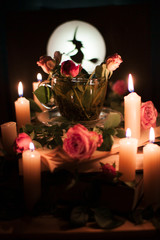 Enchanted tea surrounded by burning candles, with flowers in a cup. Silhouette of a witch on the background of a bright moon.