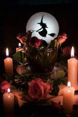 Enchanted tea surrounded by burning candles, with flowers in a cup. Silhouette of a witch on the background of a bright moon.