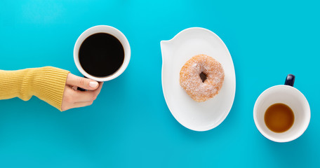 One cup of coffee held by a hand and the second one is finished, there is a doughnut on plate shaped like text bubble, placed between them, on blue background.