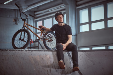 Young Bmx rider relaxing after practicing tricks leaning on his bike and looking away in a...