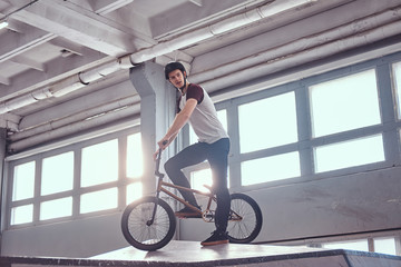 Fototapeta na wymiar Full body portrait of a young man in protective helmet with his bike standing in a skatepark indoors