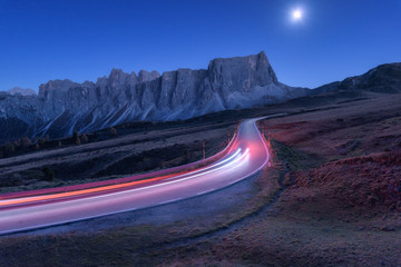 Wazig autokoplampen op kronkelende weg & 39 s nachts in de herfst. Landschap met asfaltweg, lichte paden, bergen, heuvels, blauwe lucht met maanlicht in de schemering. Rijweg in Italië. Maan over snelweg en rotsen