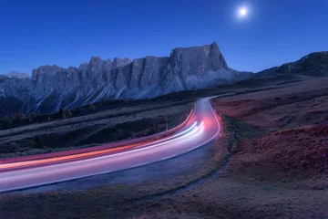 Wall murals Highway at night Blurred car headlights on winding road at night in autumn. Landscape with asphalt road, light trails, mountains, hills, blue sky with moonlight at dusk. Roadway in Italy. Moon over highway and rocks