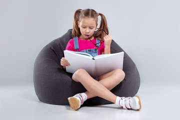 Young girl in glasses reading a book on a gray bean bag