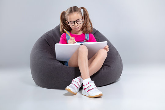 Young Girl In Glasses Doing Homework On A Gray Bean Bag