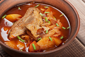 Traditional Ukrainian borsch with meat on the edge, in a brown clay bowl, on a wooden background.
