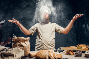 Cheerful bearded baker wearing a uniform throws up some flour standing near the table with fresh products from his bakery