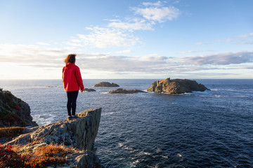 Woman in red jacket is standing at the edge of a cliff and enjoying the beautiful ocean scenery. Taken in Crow Head, North Twillingate Island, Newfoundland and Labrador, Canada. © edb3_16
