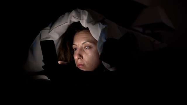 Young woman in bed with smartphone. Woman starring at cellphone device before going to bed in 4k