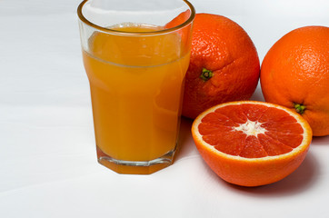 A glass of orange juice on the white background stands next to two whole oranges and sliced red orange
