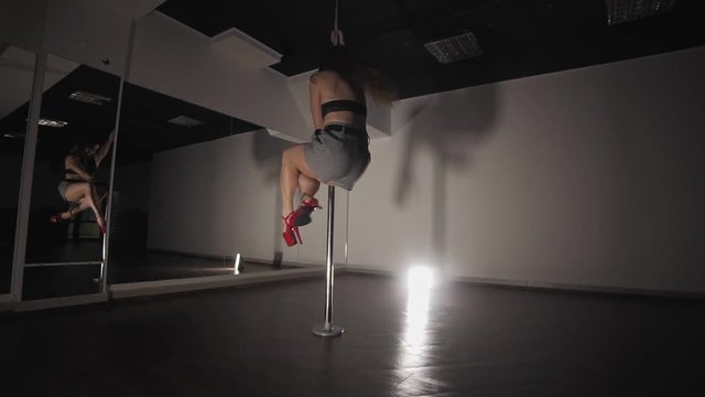 Young sexy woman pole dancing at dark studio with mirror. Woman with sexy shape is dancing near the pole in slow motion. Dancer moving with pylone