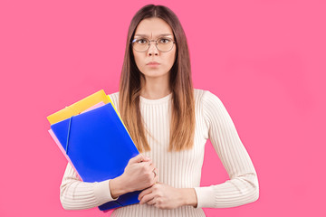 very strict girl with glasses round frame holds folders with documents in her hands on an isolated pink background