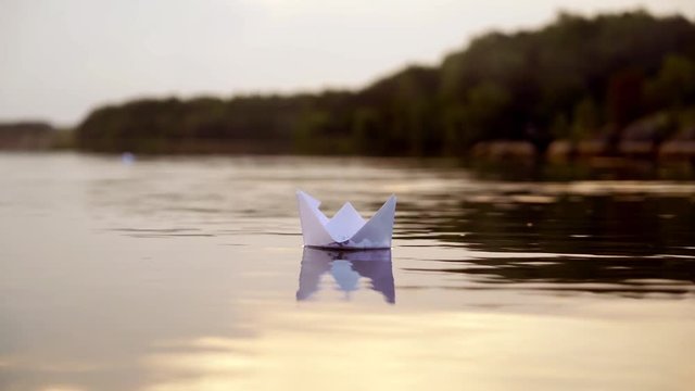 White paper boat is floating on the water near the forest background in the evening. Origami boat floats away into the distance along the river