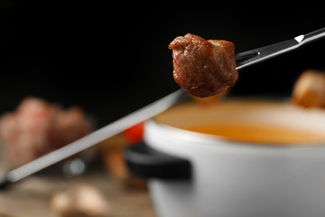 Fork with cooked meat and blurred fondue pot on background, closeup. Space for text