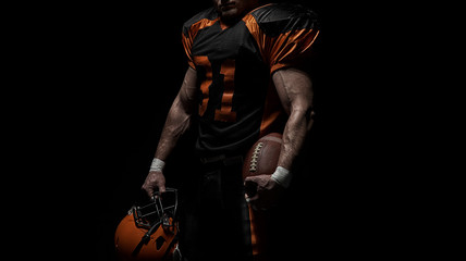 American football player on a dark background in black and orange equipment. © bondarchik
