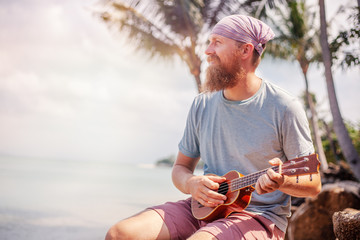 Young handsome redhead man with a beard playing a ukulele on a tropic beach, music, art, travel and vacations concept