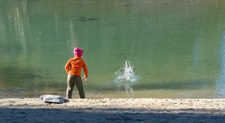 A little girl, a child, in warm clothes and a red cap throws stones into the water from the sandy...