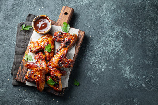 Roasted chicken wings in barbecue sauce with sesame seeds and parsley on a wooden board on a concrete table. Top view with copy space. Tasty snack for beer on a dark background. Flat lay