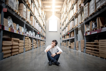 Fototapeta premium Asian shopper man sitting between cardboard box shelves aisle in warehouse choosing what to buy. Shopping lifestyle in department store. Buying or purchasing factory goods. Inventory industry concept