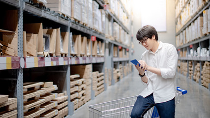 Asian man shopper standing with trolley cart in warehouse inventory aisle checking his shopping...