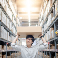 Asian delivery man carrying cardboard box overhead in product shelves aisle in warehouse. Inventory picking and packing. Shopping lifestyle in department store. Buying or purchasing factory goods
