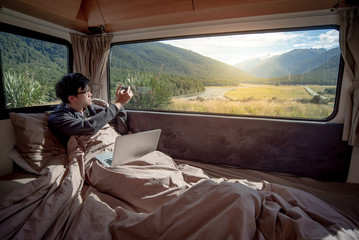 Asian man traveler enjoy taking photo of mountain scenery through the window by smartphone while staying in the blanket in camper van in the morning. Road trip in summer of South Island, New Zealand. - obrazy, fototapety, plakaty