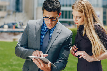 Handsome businessman in a city. Businessman in a glasses. Business couple in a summer city. Man and woman standing with tablet. Pretty blonde in a dress with her parther.