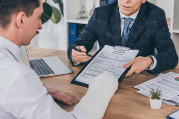 businessman in blue jacket giving form for compensation claim to worker with broken arm at table in office, compensation concept - obrazy, fototapety, plakaty