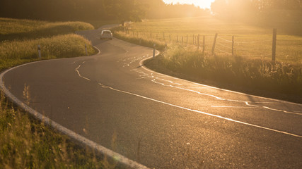 Zigzagging serpentine road between meadows just before sundown metaphorically difficult way. Landscape backlit warm colored colorful scene.