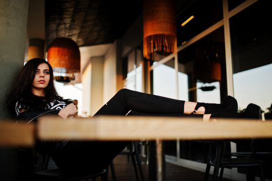 Young Curly Woman In Leather Jacket In A Bar Put Her Legs On Table.