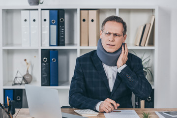 businessman in neck brace sitting at table, feeling pain and holding neck in office, compensation concept