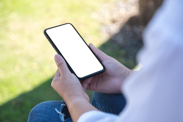 Mockup image of a woman holding black mobile phone with blank white screen while sitting in the outdoors