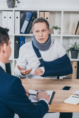 Fototapeta premium middle aged worker in neck brace with broken arm sitting at table and talking to opposite businessman in office, compensation concept
