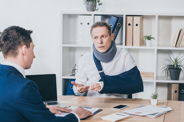 middle aged worker in neck brace with broken arm sitting at table and talking to businessman in blue jacket in office, compensation concept - obrazy, fototapety, plakaty