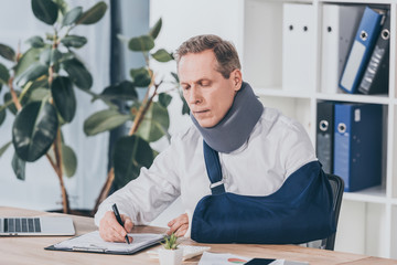 worker in neck brace and arm bandage sitting at table and carefully writing on document with pen in office, compensation concept - obrazy, fototapety, plakaty