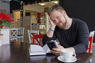Cheerful bearded man in black shirt drinking espresso, looking at camera and chatting with friends social network apps on his smartphone. handsome man freelancer is sitting at the table of the coffee 