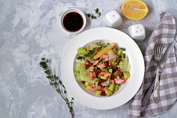 Green salad with avocado, baked salmon and dried tomatoes in a white plate on a gray concrete background. Top view.