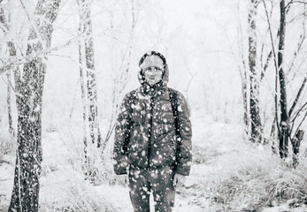 Fototapeta na wymiar A guy wearing a jacket with a hood is standing in a snowy forest, snow is falling on him from above. Black and white photo